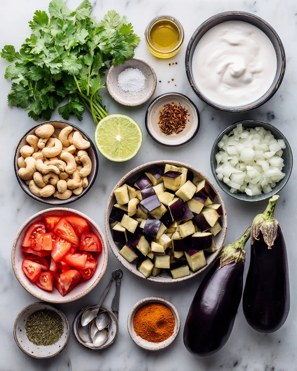 Flat lay of creamy eggplant curry ingredients: eggplant, onion, garlic, ginger, coconut milk, tomatoes, spices, cashews, cilantro, lime.