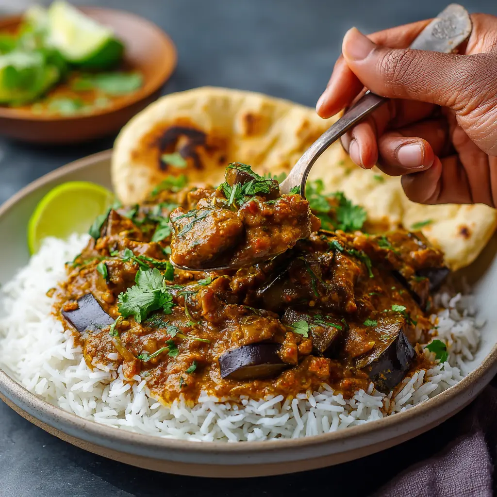 Spoon serving creamy eggplant curry over basmati rice on a white plate, with naan and lime; steam rising, cozy and inviting.