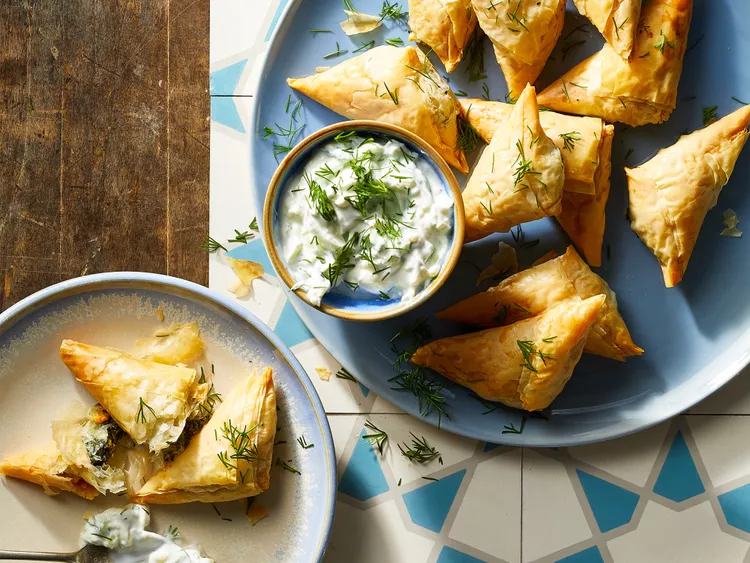 Golden brown Air Fryer Spanakopitas arranged neatly on a white serving plate, garnished with chopped fresh dill. Each piece shows crispy, flaky layers of phyllo pastry filled with a vibrant green spinach and cheese mixture, accompanied by a small bowl of creamy tzatziki sauce on the side.