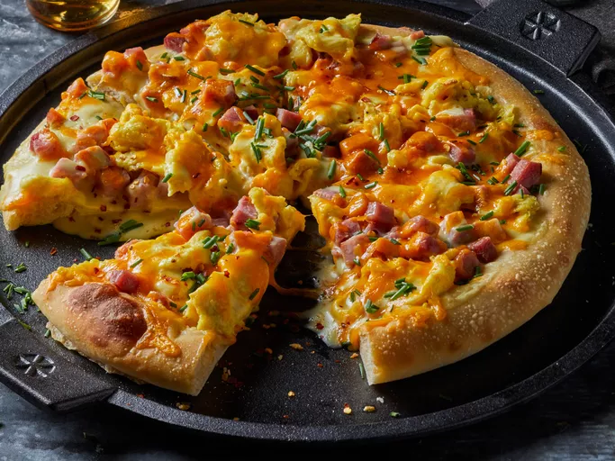 A freshly baked Eggs Benedict Breakfast Pizza on a wooden table, topped with creamy Hollandaise sauce, scrambled eggs, diced ham, and melted cheese