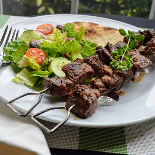 Grilled Lamb Souvlaki skewers on a platter, served with rosemary garlic roasted potatoes, Greek salad, and pita bread on a rustic wooden table