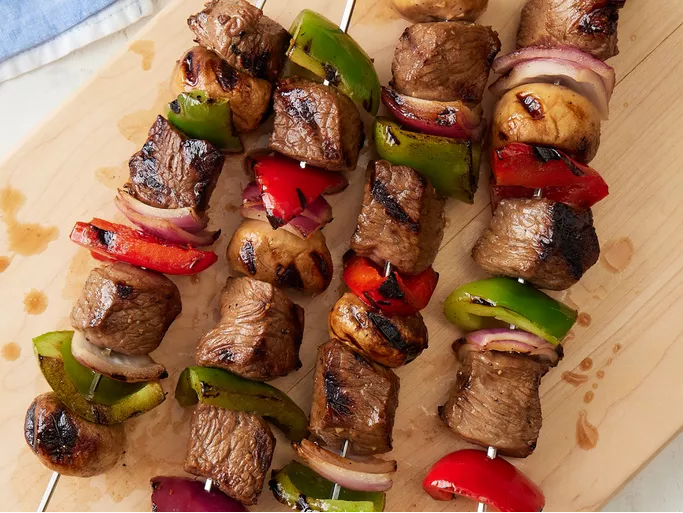 Grilled beef shish kabobs with mushrooms, green and red bell peppers, and onions on a skewer, served on a white plate.