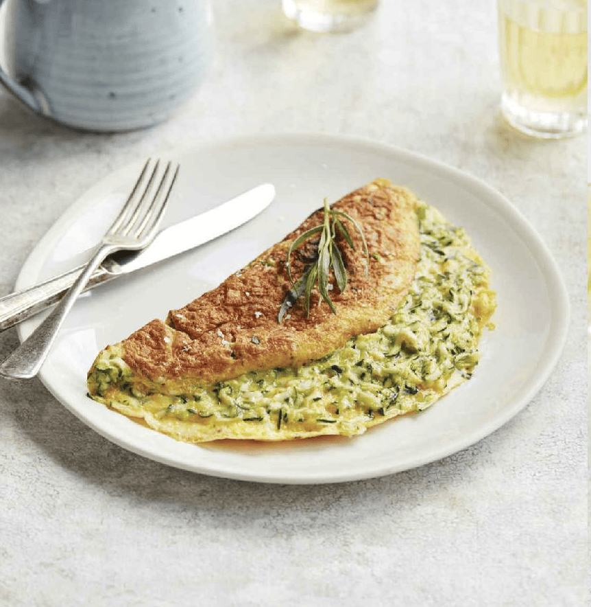 A fluffy zucchini omelet served on a white plate, garnished with fresh herbs and slices of zucchini. The omelet is golden brown and lightly crisped on the edges, with vibrant green zucchini pieces visible throughout