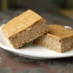 Stack of homemade high-fiber, high-protein breakfast bars on a white plate, topped with a drizzle of natural peanut butter.