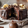 Moist chocolate banana cake on a stand with glossy ganache drips and a slice removed showing soft, tender crumb