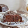 Moist chocolate banana cake on a stand with glossy ganache drips and a slice removed showing soft, tender crumb