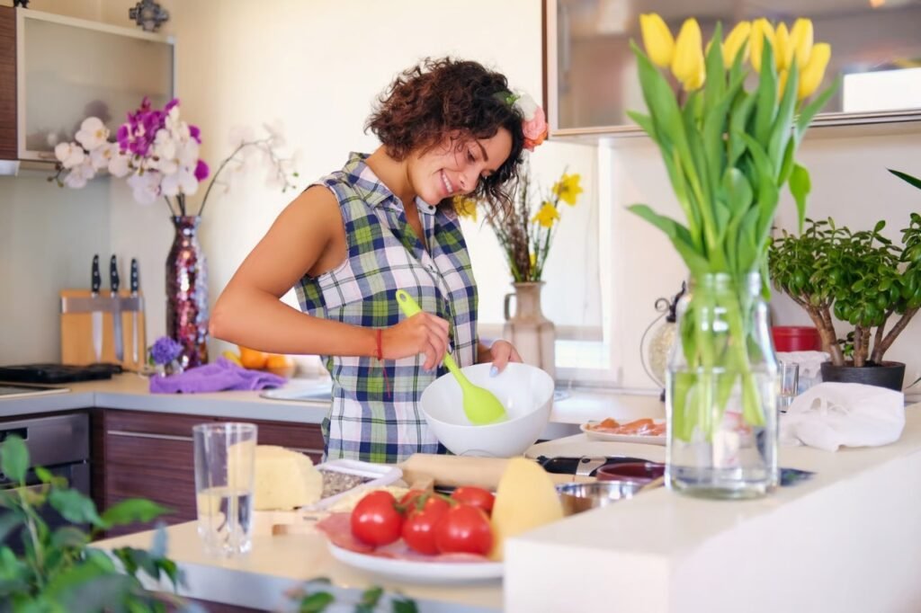 Alissa Savoldelli smiling while mixing ingredients in a white bowl in her bright kitchen, surrounded by fresh vegetables, flowers, and cooking tools.