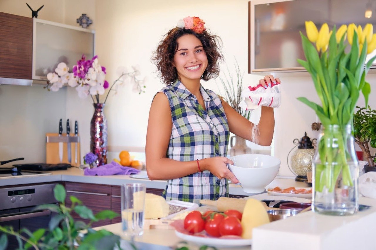 Alissa Recipes smiling in her kitchen while preparing ingredients for a recipe, surrounded by fresh vegetables and flowers.